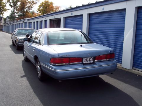 2001 Ford Crown Victoria - A light blue Ford Crown Victoria is parked in front of a row of blue storage unit doors. Another car is behind it.
