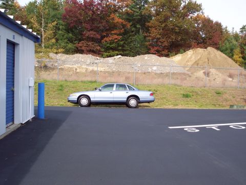 2001 Ford Crown Victoria - A light blue 2001 Ford Crown Victoria sedan is parked next to a storage unit with a wooded area and dirt piles in the background.