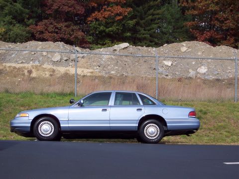 2001 Ford Crown Victoria - A light blue 2001 Ford Crown Victoria sedan is seen from the side, parked next to a road with a chain-link fence and wooded area in the bac…