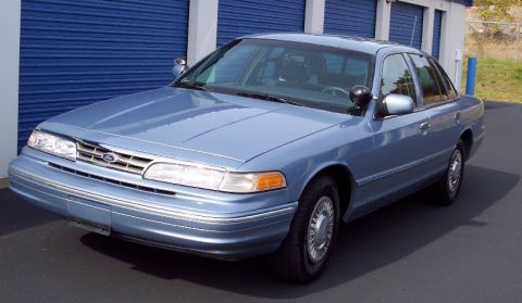 2001 Ford Crown Victoria - This light blue sedan is parked outside storage units. It appears to be a police vehicle.