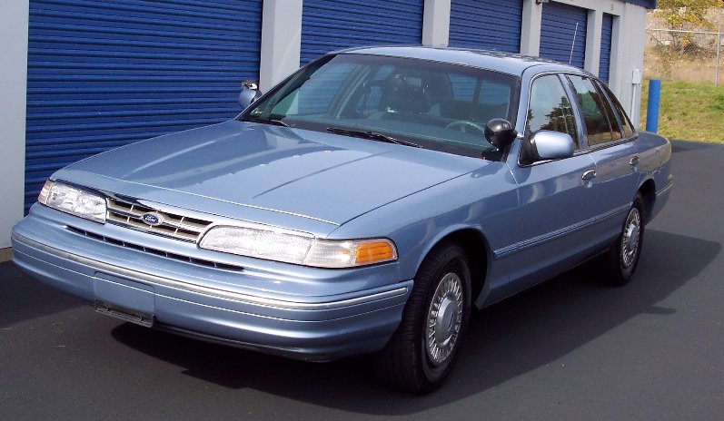 Light blue Ford Crown Victoria parked in front of blue storage unit doors. Car has a police spotlight on the driver's side mirror