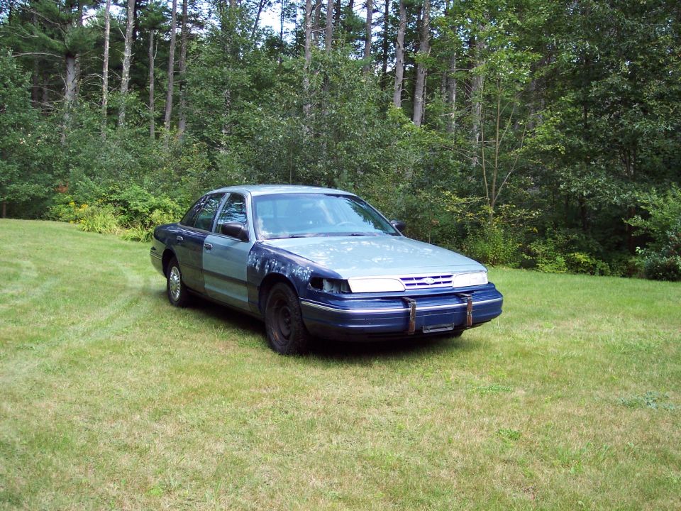Two-tone blue 1990s Ford Crown Victoria with missing headlight parked on grass