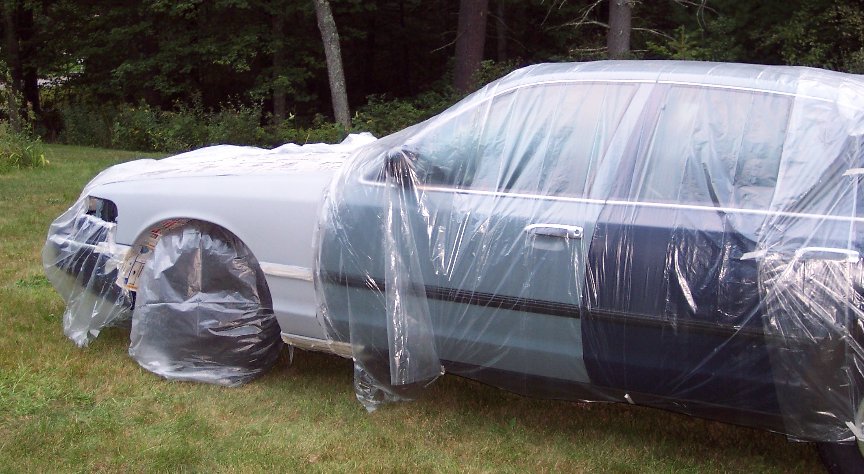 Side view of a 2001 Ford Crown Victoria covered in plastic sheeting, parked on grass