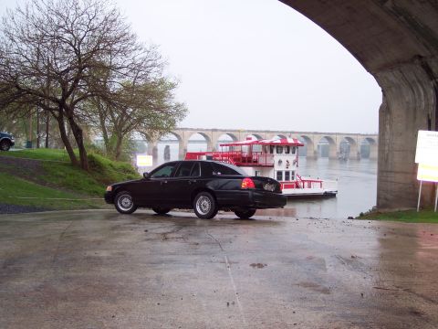 2001 Ford Crown Victoria - A black 2001 Ford Crown Victoria is parked on a wet surface near a river, with a paddleboat and a bridge in the background.