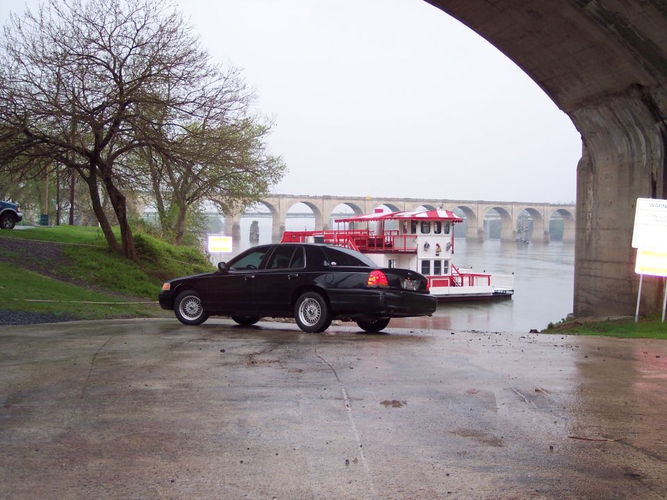Black Crown Victoria parked on a wet boat ramp near a red paddlewheel riverboat