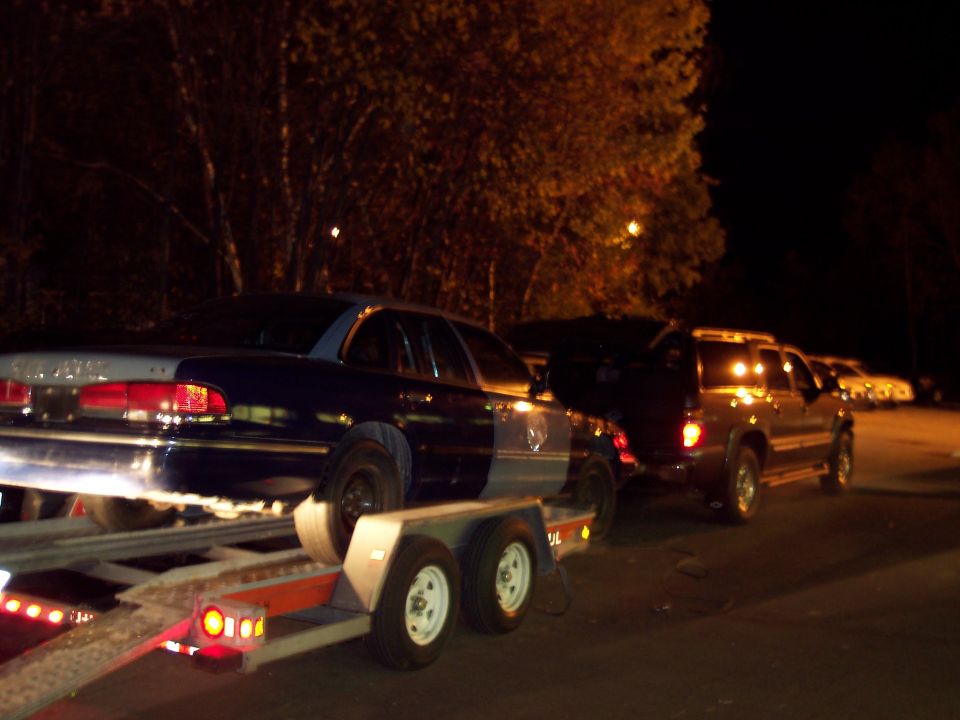 Dark SUV at night towing a car trailer carrying a light blue sedan