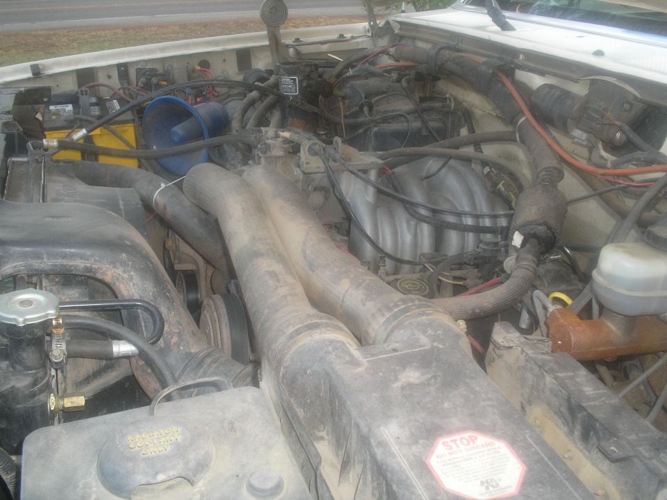 Engine bay with V8 engine, intake manifold, battery, and hoses visible