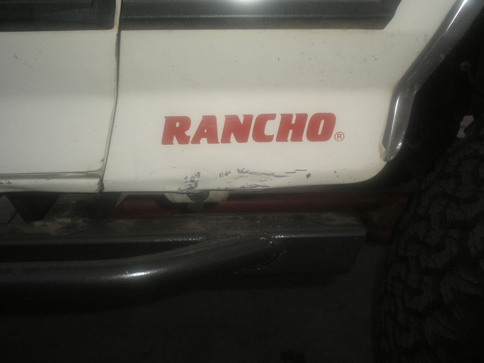 White SUV side panel with red "RANCHO" decal and scuffs, black side step, tire visible