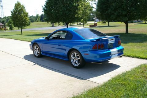 1998 Ford Mustang - A blue 1998 Ford Mustang coupe with white racing stripes is parked on a concrete driveway.