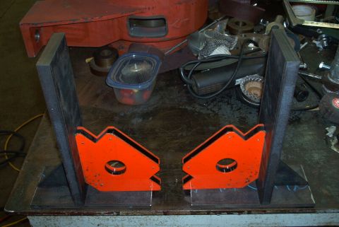 1983 Ford Bronco - Orange welding magnets and metal brackets are arranged on a workbench with various tools and parts in the background.