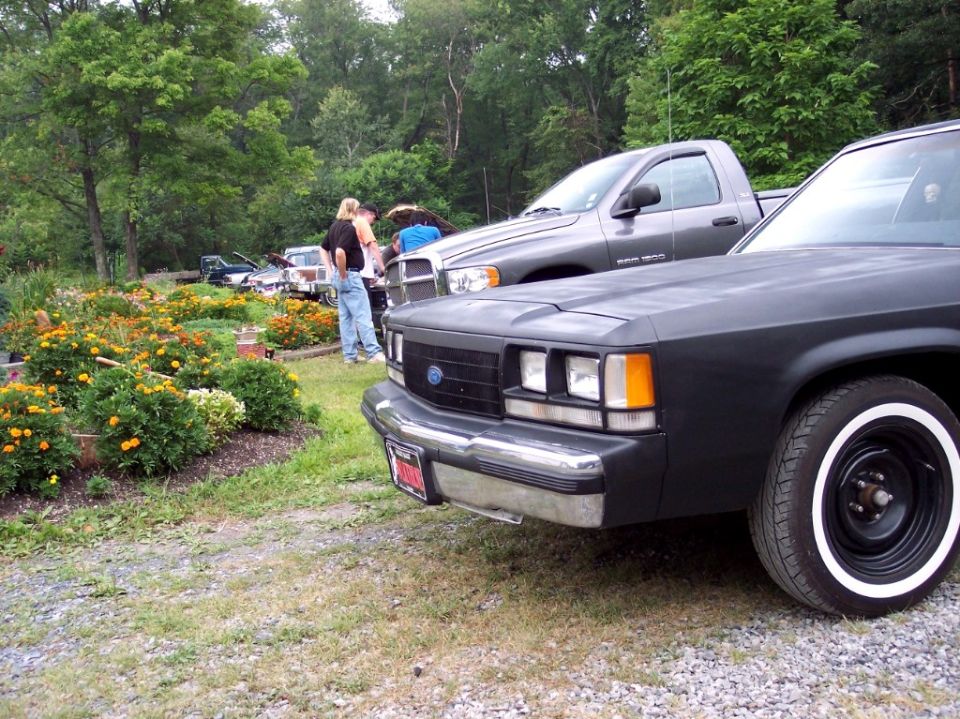 Flat-black Ford sedan front, chrome bumper, whitewall tires, parked on gravel by flowers