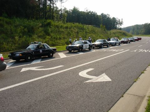 1993 Ford Mustang - A convoy of police vehicles is parked along the road, with a few people standing nearby.