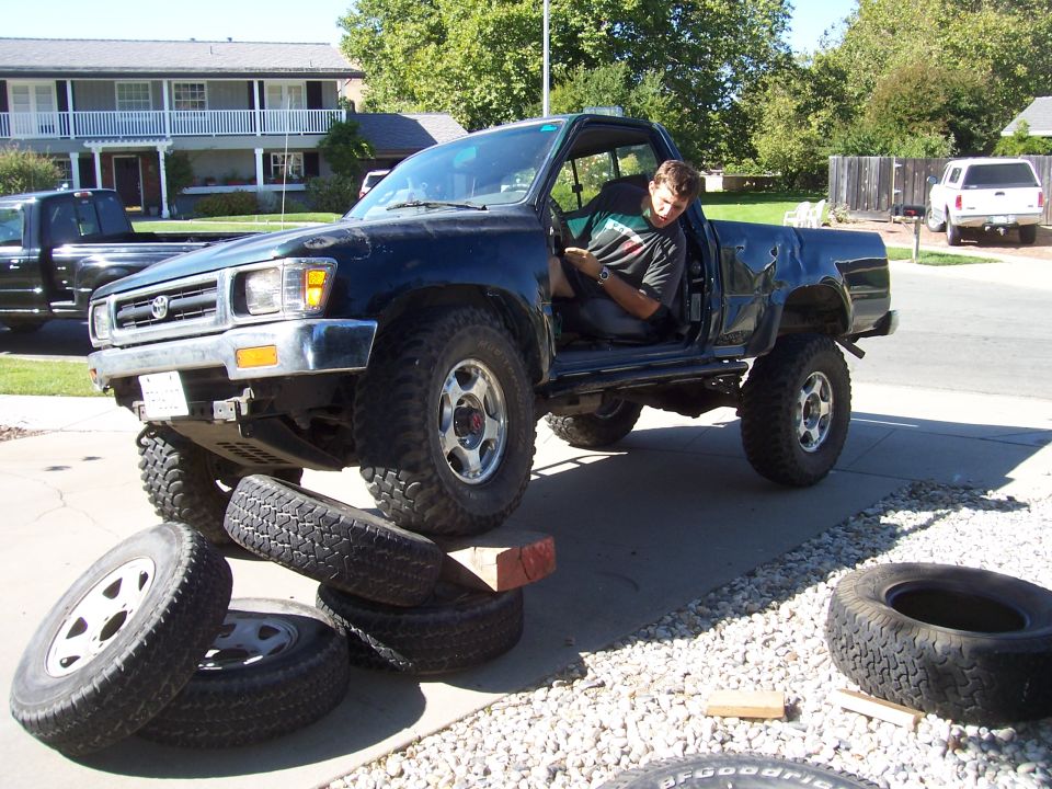 Green Toyota pickup with front wheels perched on a stack of tires and lumber in a driveway, man in doorway