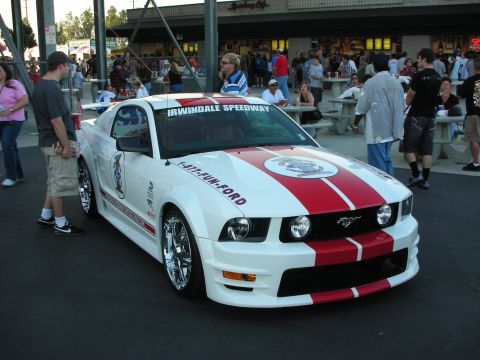 1998 Ford Crown Victoria - A white Ford Mustang with red racing stripes and various decals is parked outside a building with people around.