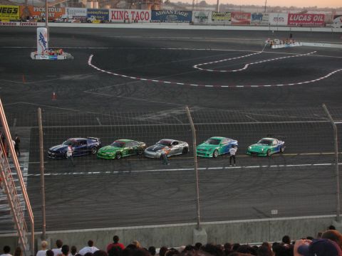 1998 Ford Crown Victoria - A group of tuned cars are positioned on a race track, with spectators visible in the lower portion of the frame.
