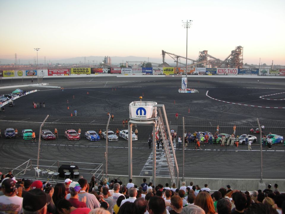 Cars lined up at dusk on a paved oval drift track with grandstand crowd