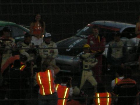1998 Ford Crown Victoria - Race cars and crew members are visible behind a fence at what appears to be a racing event.