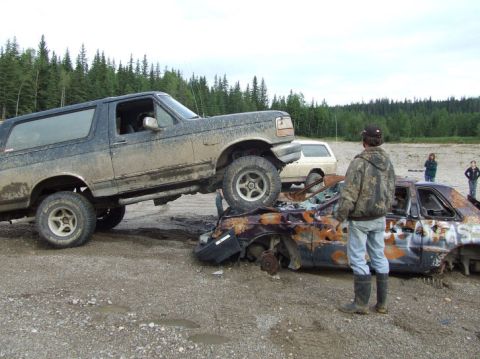 1988 Ford Bronco - A muddy Ford Bronco is positioned on top of a heavily damaged car in an outdoor area.