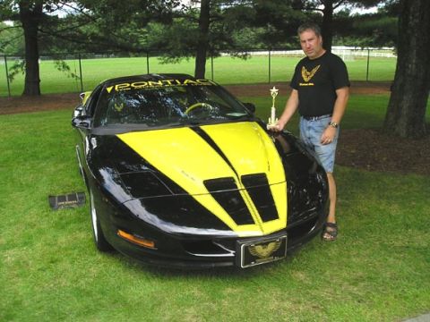 1984 Pontiac Fiero - A man holds a trophy next to a black and yellow 1994 Pontiac Trans Am with racing stripes.