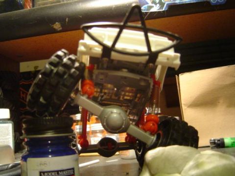 1979 Ford Bronco - Underside view of a partially built scale model truck on a workbench, showing the chassis with a solid front axle, orange coil-over shocks,…