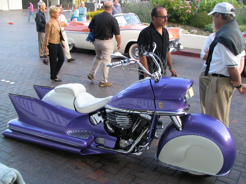 Custom purple motorcycle with white seats and large fins, parked on brick pavement. Two classic cars and people are in the background