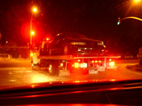 1993 Ford Taurus - A black sedan is being towed on a flatbed truck at night, illuminated by red and yellow lights.