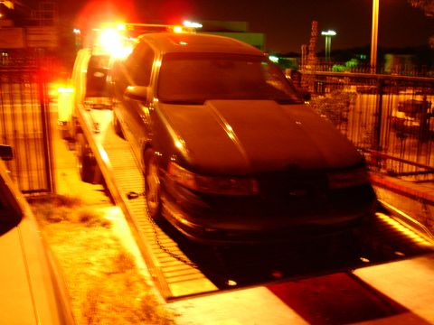 1993 Ford Taurus - A dark 1993 Ford Taurus is loaded onto a tow truck at night, its hazard lights flashing.
