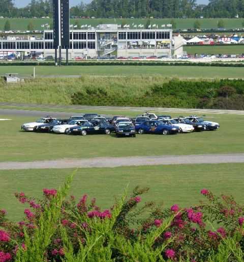 1993 Ford Mustang - A collection of vehicles, including police cruisers, are gathered on a grassy field with a race track and grandstand visible.