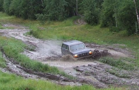 1988 Ford Bronco - Dark full-size Ford Bronco with headlights on blasting through a wide muddy water hole, throwing a heavy spray of brown water off its passe…