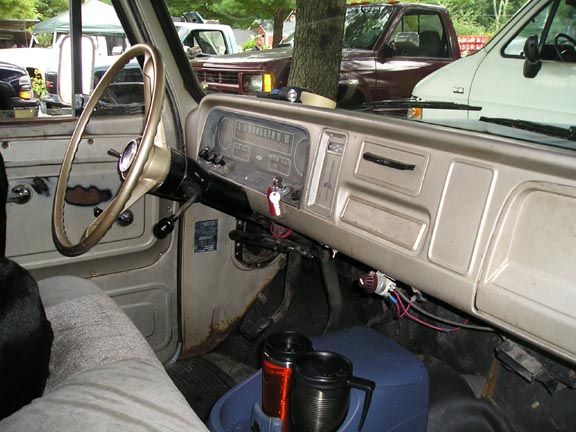 Old truck interior showing dashboard, steering wheel, and gauges. Other vehicles visible outside