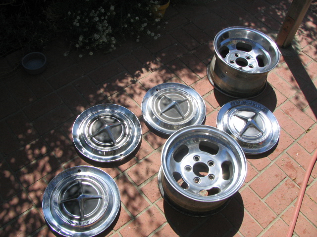 Two polished aluminum slot mag wheels and three chrome hubcaps arranged on a red brick patio