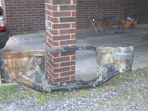 1978 Ford Bronco - A heavily rusted vehicle radiator support is shown resting on the ground next to a brick pillar.
