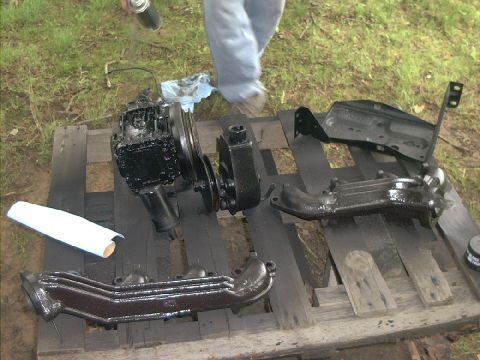 1978 Ford Bronco - Engine parts are being painted black outdoors on wooden pallets.