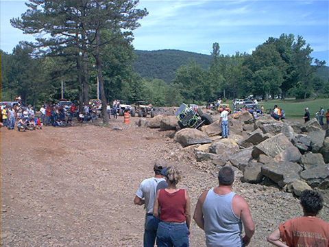 1978 Ford Bronco - An off-road vehicle is stuck on a pile of rocks, surrounded by onlookers at an outdoor event.