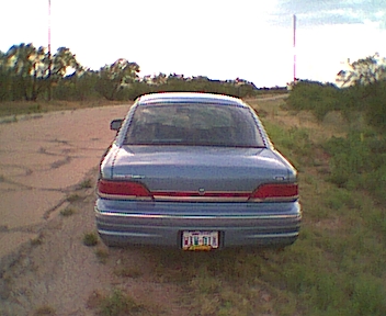 1994 Ford Crown Victoria - The rear of a blue sedan is shown parked on a dirt road with vegetation on either side.