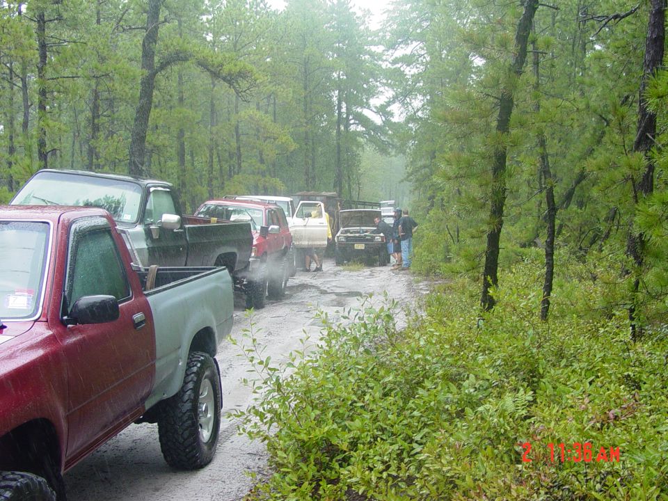 Line of off-road vehicles on a muddy dirt road in a forest, some with hoods open
