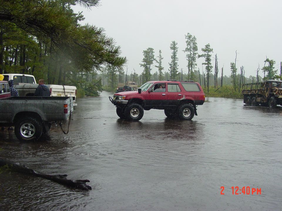Red SUV with lifted suspension driving through water, other trucks visible