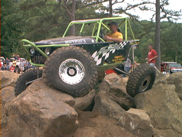 Black off-road buggy with green roll cage climbing rocks, large tires, and 'Monroes' text