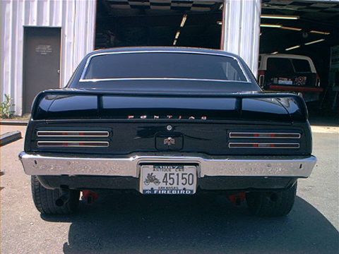 1978 Ford Bronco - Rear view of a black Pontiac Firebird, showing its distinctive taillights and spoiler, with another vehicle visible in the background.