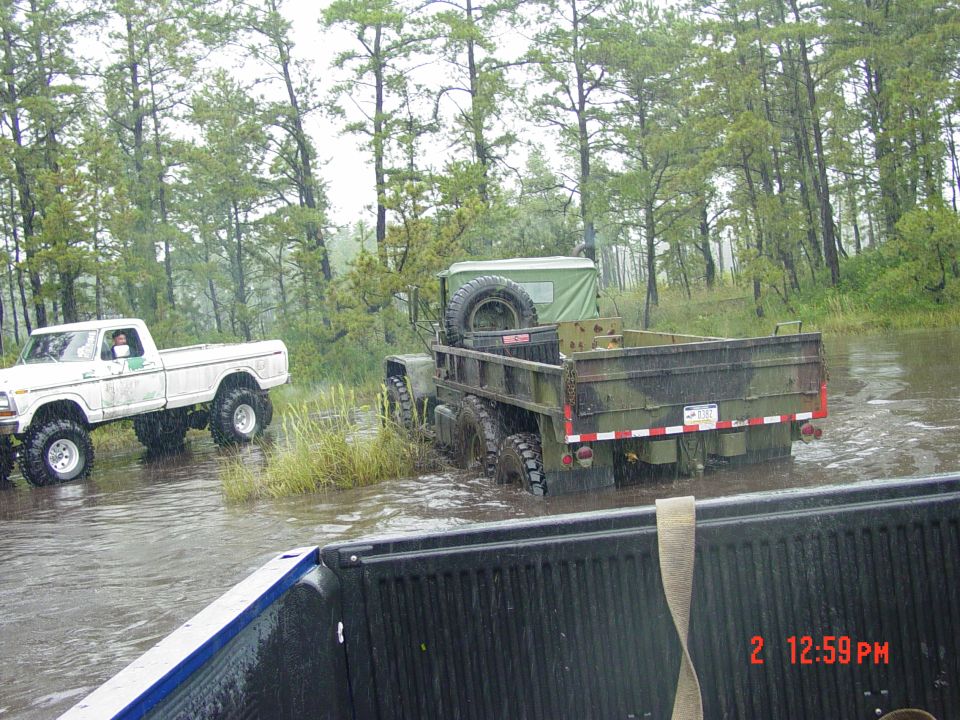 Rear view of a camouflaged military truck driving through water, with a white pickup truck behind it