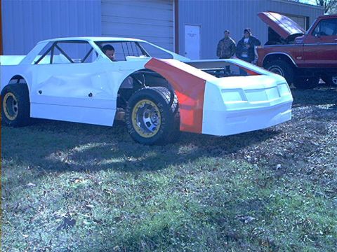 1978 Ford Bronco - A race car project is parked outside a building with a red pickup truck behind it.