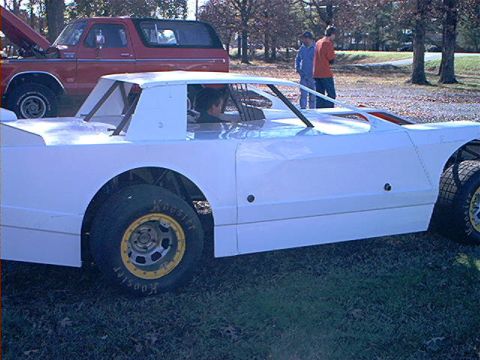 1978 Ford Bronco - A white stock car is parked outdoors next to a red pickup truck with its hood open, with a few people standing in the background.