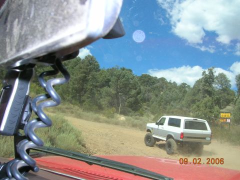 1985 Ford Bronco - A white 1985 Ford Bronco is seen from behind as it drives on a dusty dirt road through a wooded area.