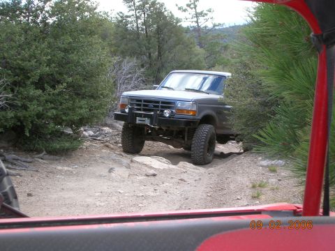 1985 Ford Bronco - Gray Ford Bronco navigates a rocky off-road trail surrounded by trees and brush.