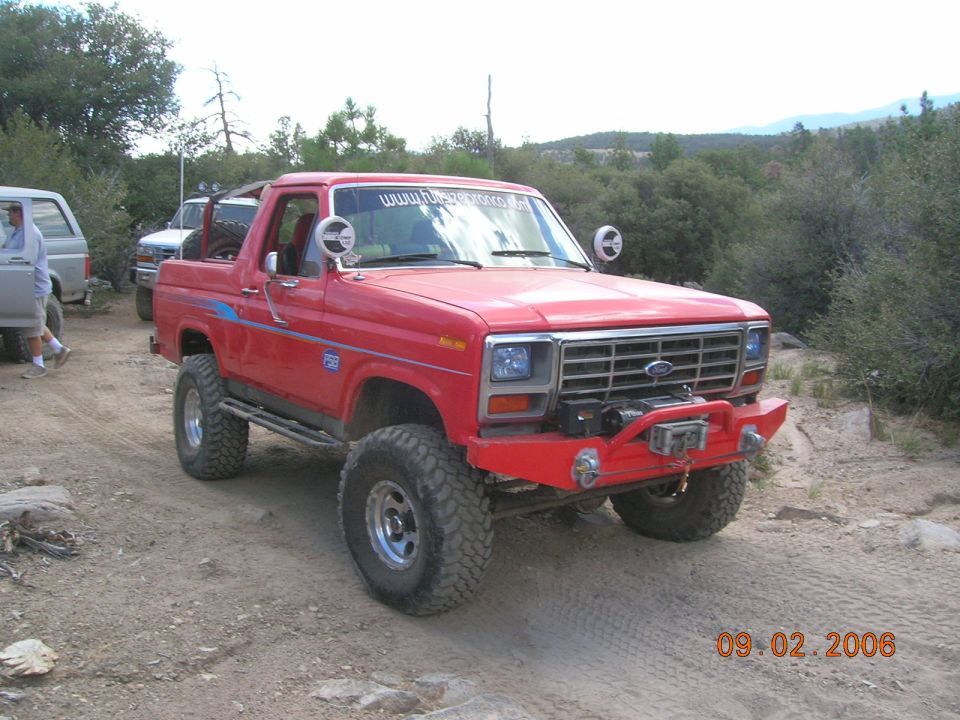 Red Ford Bronco with lifted suspension and off-road tires parked on a dirt trail. Another SUV is visible to the left