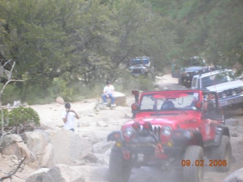 1985 Ford Bronco - Several off-road vehicles navigate a rocky trail surrounded by trees and brush.