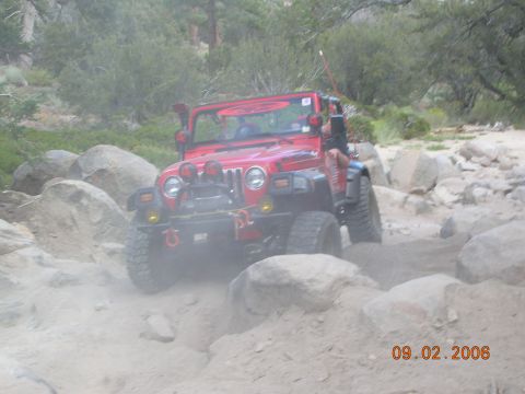1985 Ford Bronco - Red Jeep Wrangler tackling a challenging off-road course with large rocks.