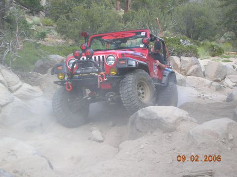 1985 Ford Bronco - A red Jeep Wrangler navigates a rocky off-road trail, kicking up dust.