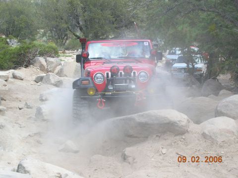 1985 Ford Bronco - A red Jeep Wrangler navigates a dusty, rocky off-road path with other vehicles in the background.