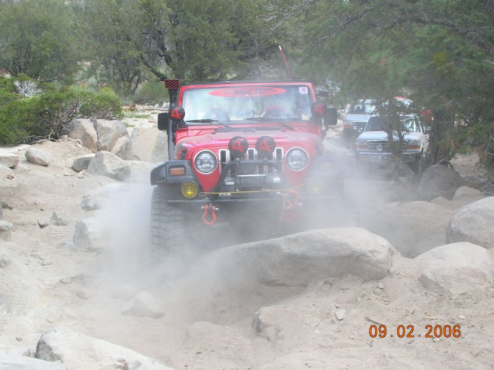 Red Jeep Wrangler driving through dust on a rocky trail, with other vehicles behind